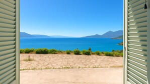 Une vue à travers une fenêtre à volets, dévoilant une terrasse en pierre, une mer d’un bleu profond et des montagnes lointaines.