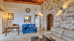 A living room with a stone wall, a wooden ceiling, a blue sofa, a beige sofa, a wooden table, and a clock on the wall. - Filistron Seaview Bliss - Coastal Charm Hideaway (Taktikoupoli)