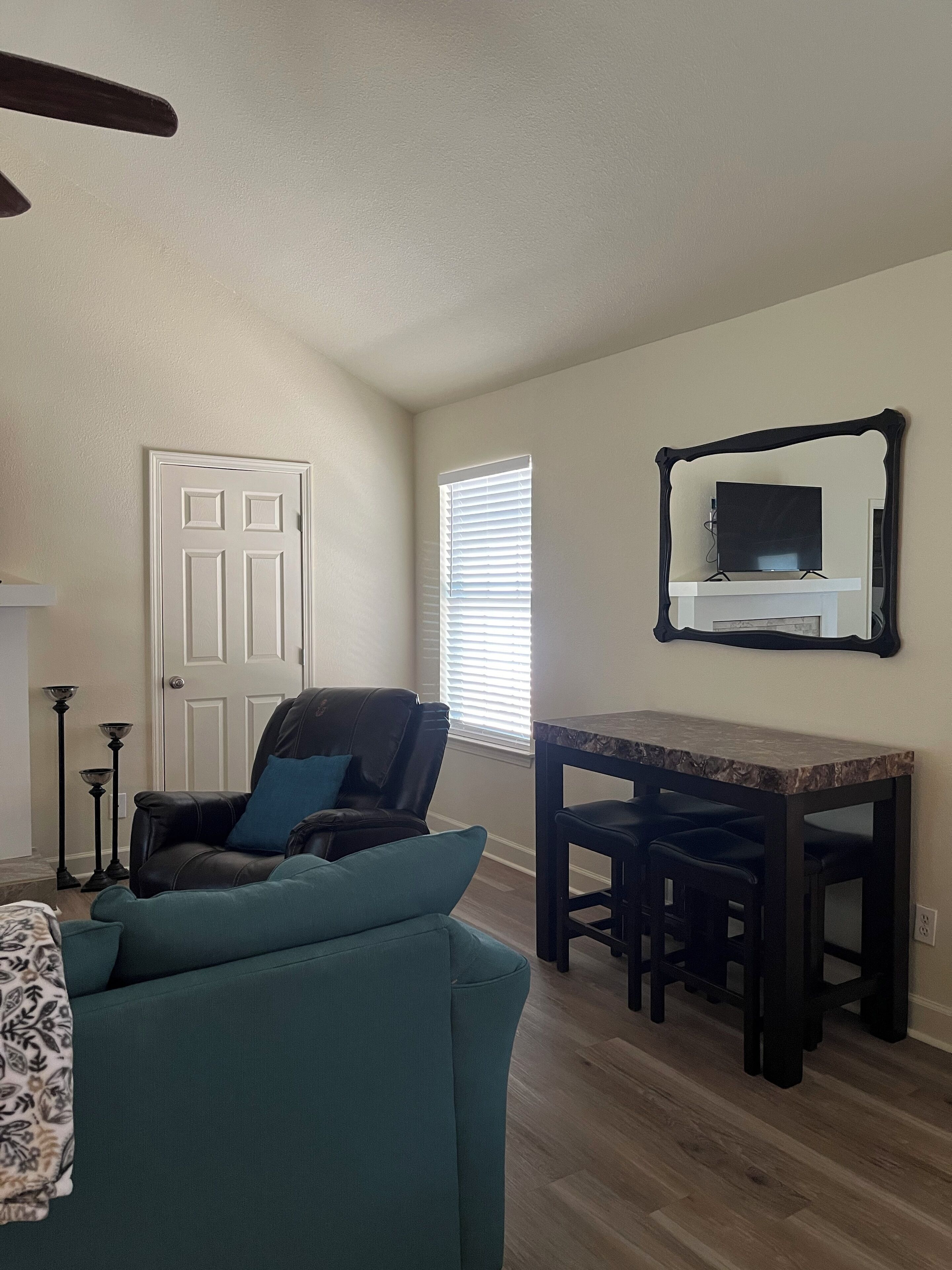 A living room with a teal sofa, a black leather chair, a wooden dining table with two black barstools, a white door, and a window with blinds.
