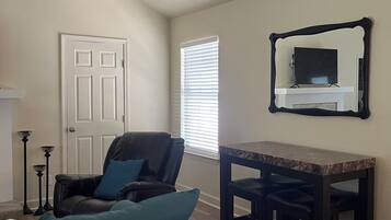 A living room with a teal sofa, a black leather chair, a wooden dining table with two black barstools, a white door, and a window with blinds.