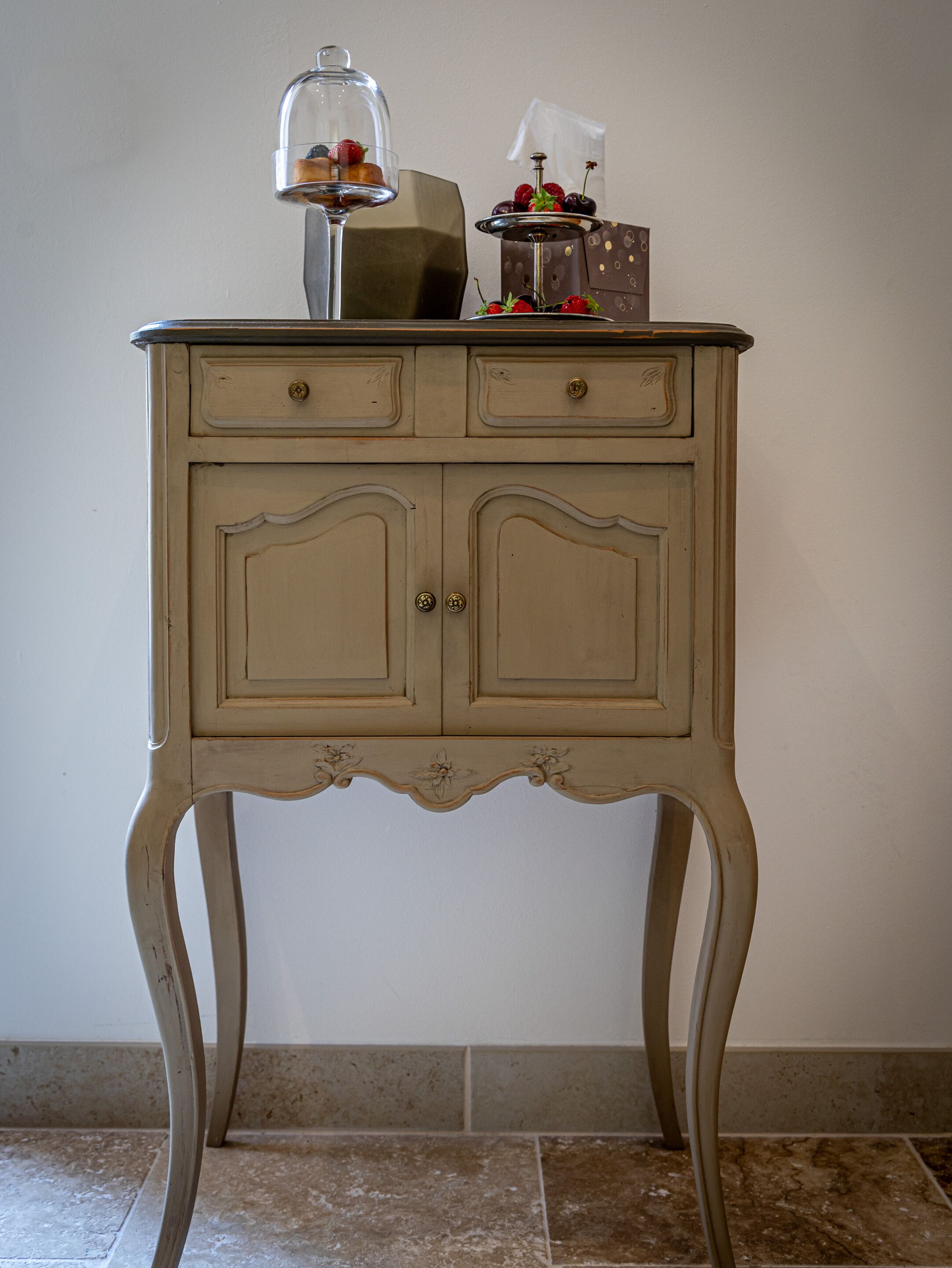 Una mesa de estilo vintage con dos cajones y una cúpula de cristal que exhibe frutas y una copa de vino.