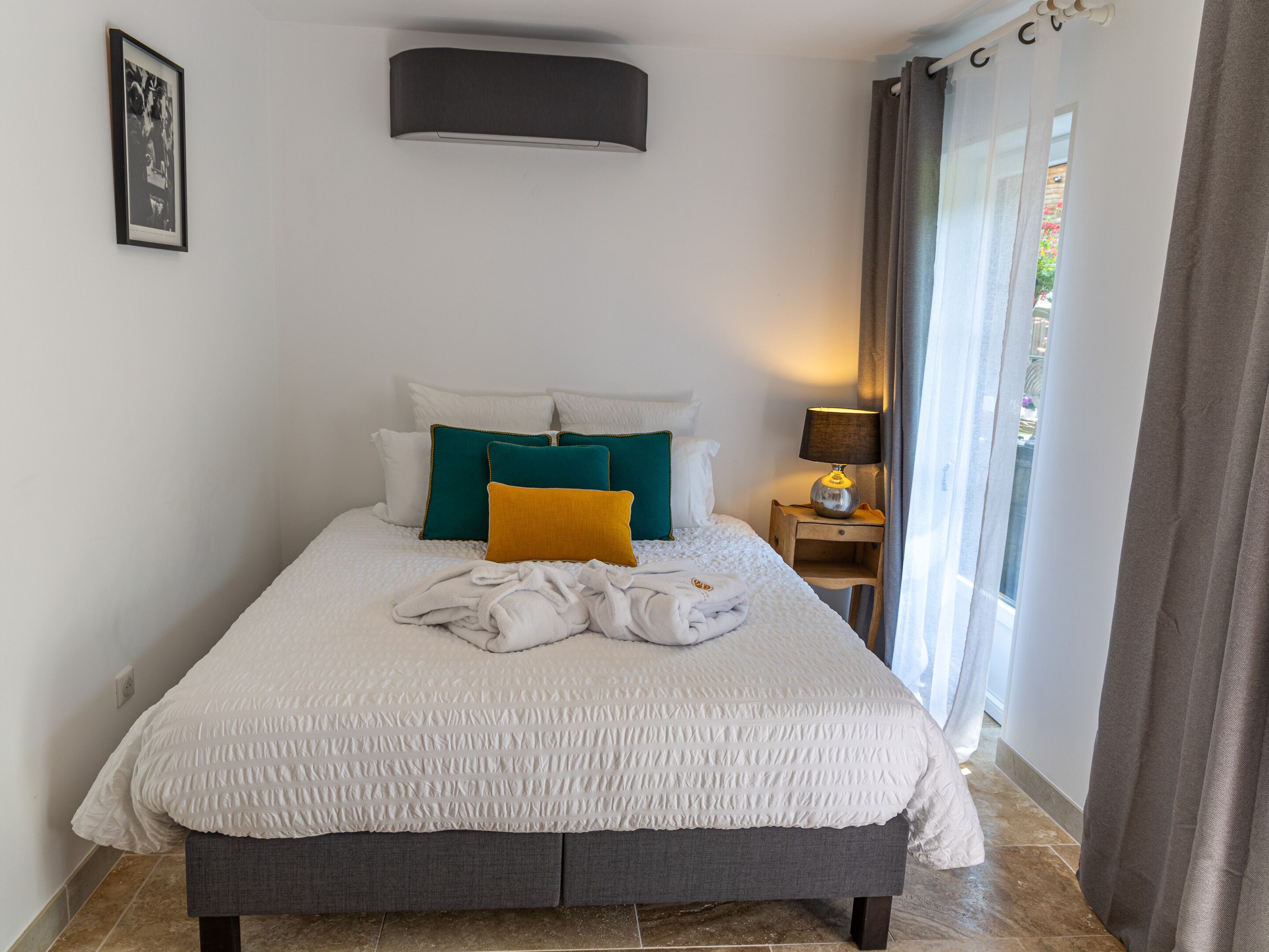 Un lit bien fait, avec une couette blanche, des coussins bleu sarcelle et moutarde, et un banc gris au pied du lit.