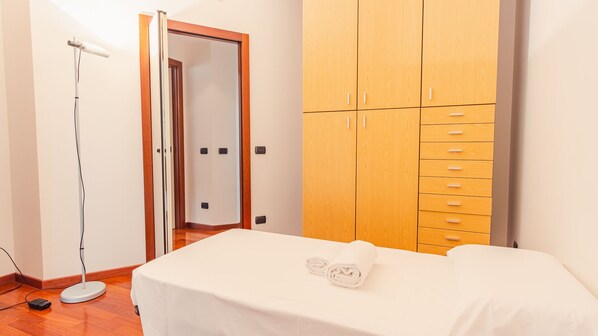 A bedroom with a bed, a wooden wardrobe, a floor lamp, and a door leading to another room.