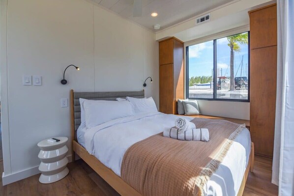 Una cama bien hecha con cabecera de madera, una lámpara de noche y vistas al exterior a través de un ventanal grande.