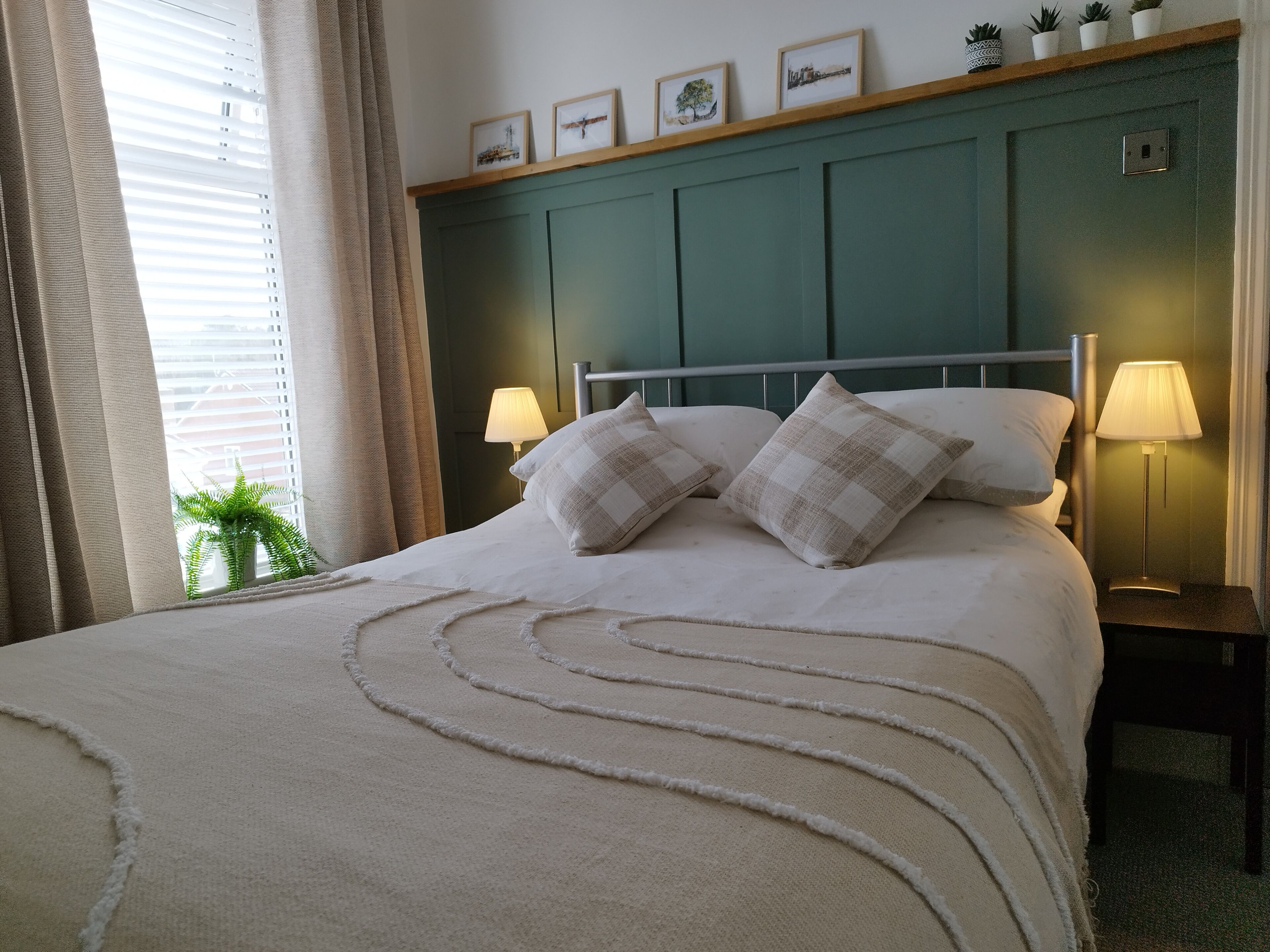 A neatly made bed with a checkered throw, a bedside lamp, and a wall-mounted shelf with framed pictures.