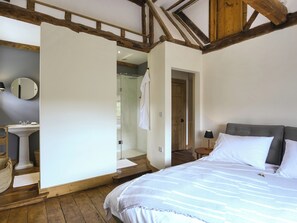 A bedroom with a bed, a bedside table with a lamp, a wooden floor, a white wall, and a wooden ceiling beam.
