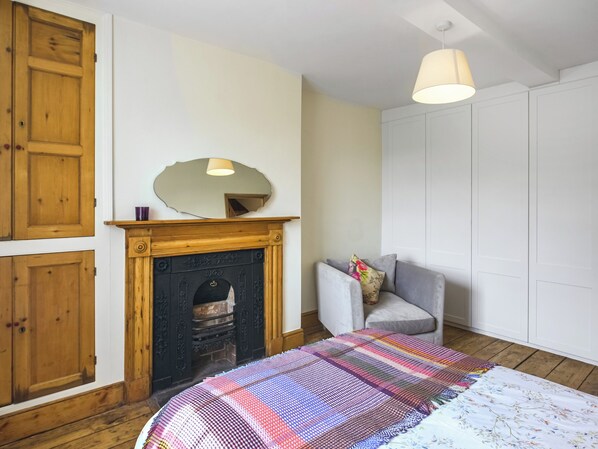 A bedroom with a fireplace, a bed with a colorful quilt, a grey armchair, and a white wardrobe.