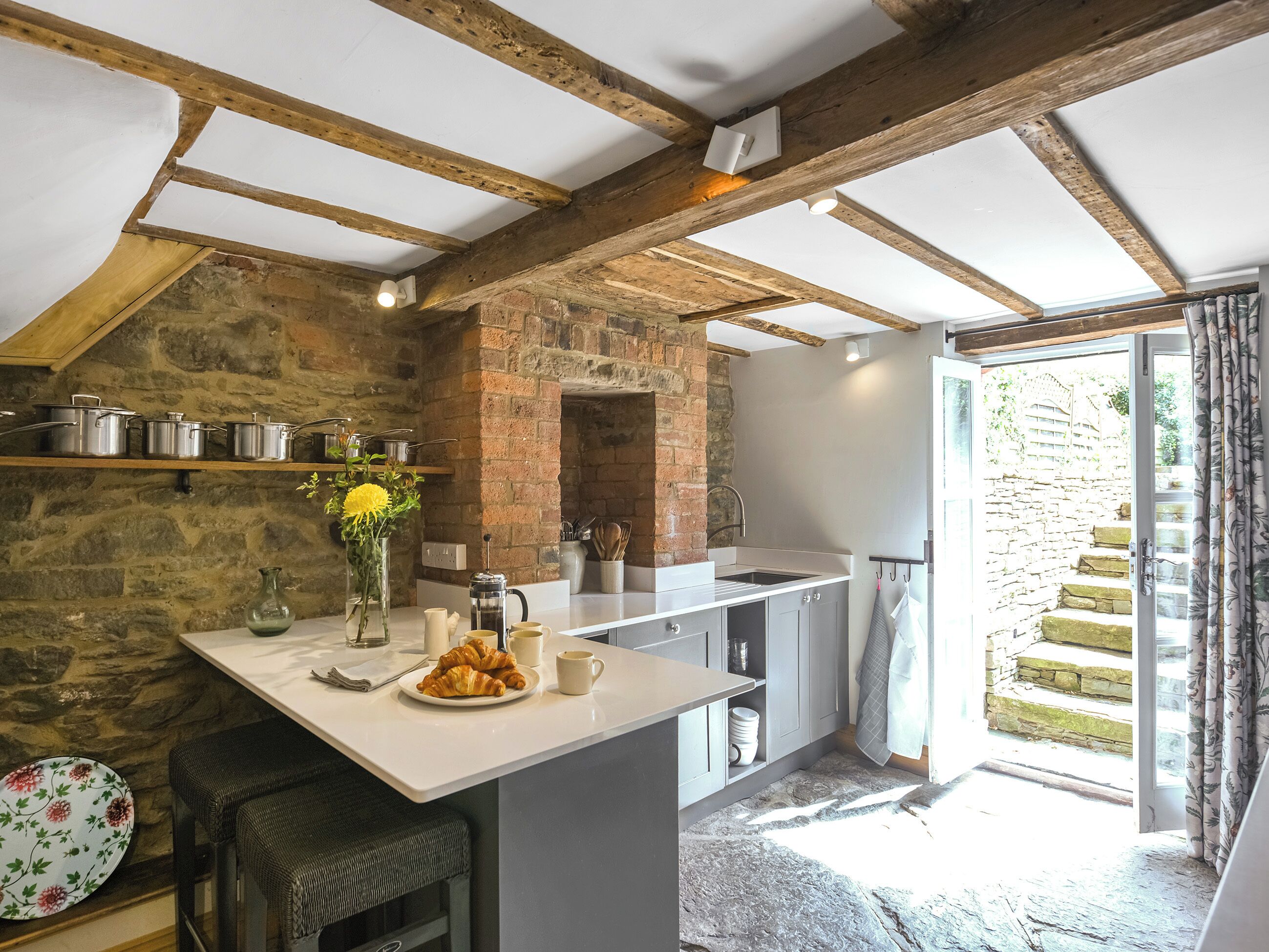 A kitchen with exposed wooden beams, stone walls, and a view of a stone staircase outside.