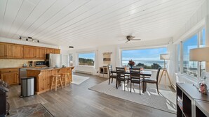 A spacious kitchen with wooden cabinets, a central island with stools, a dining area with a table and chairs, and large windows offering ocean views. - Treasure Hideaway-Manzanita Or., Ocean View,Fenced Yard, Arcade games, Dog Ok (Nehalem)