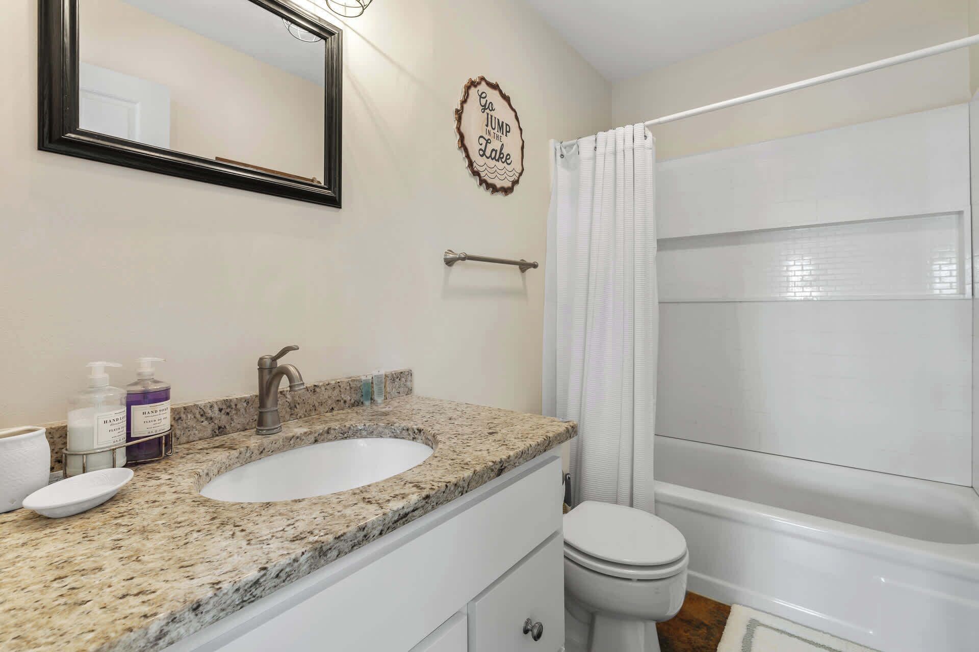 A bathroom with a double sink, a toilet, a shower curtain, and a decorative sign on the wall.