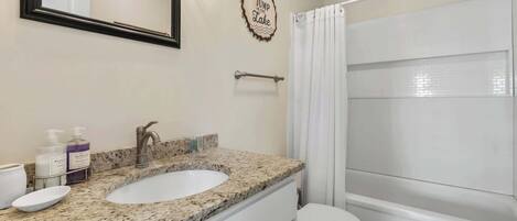 A bathroom with a double sink, a toilet, a shower curtain, and a decorative sign on the wall.