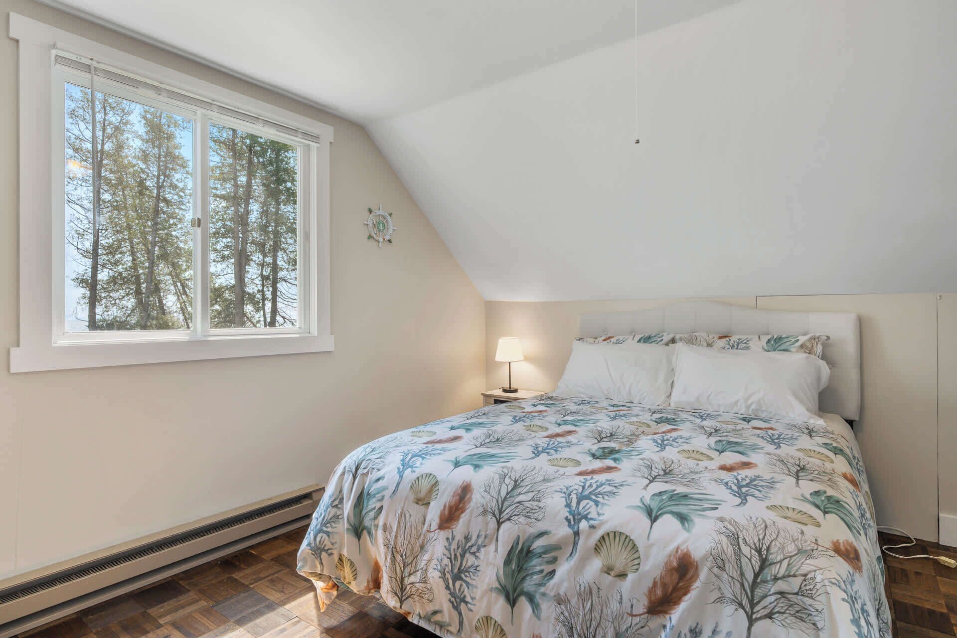 A bedroom with a large window, a bed with floral bedding, a bedside table with a lamp, and a wall-mounted clock.