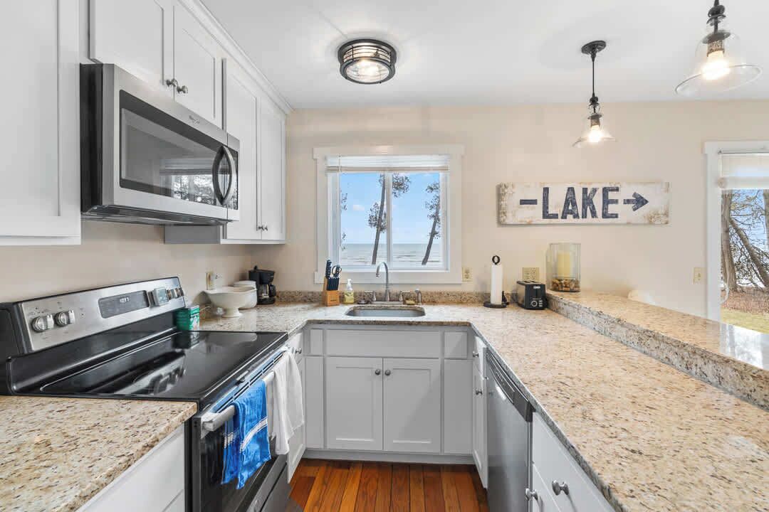 Una cocina con gabinetes blancos, un microondas de acero inoxidable, un horno doble, encimeras de granito, una ventana con vistas a árboles y un cartel que dice 'LAKE'.