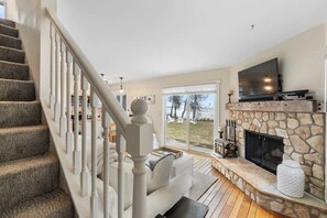 A living room with a stone fireplace, a large flat-screen TV, a staircase, and a view of the outdoors. - Waterfront Lake Michigan Tranquility Bay Getaway (Sister Bay)