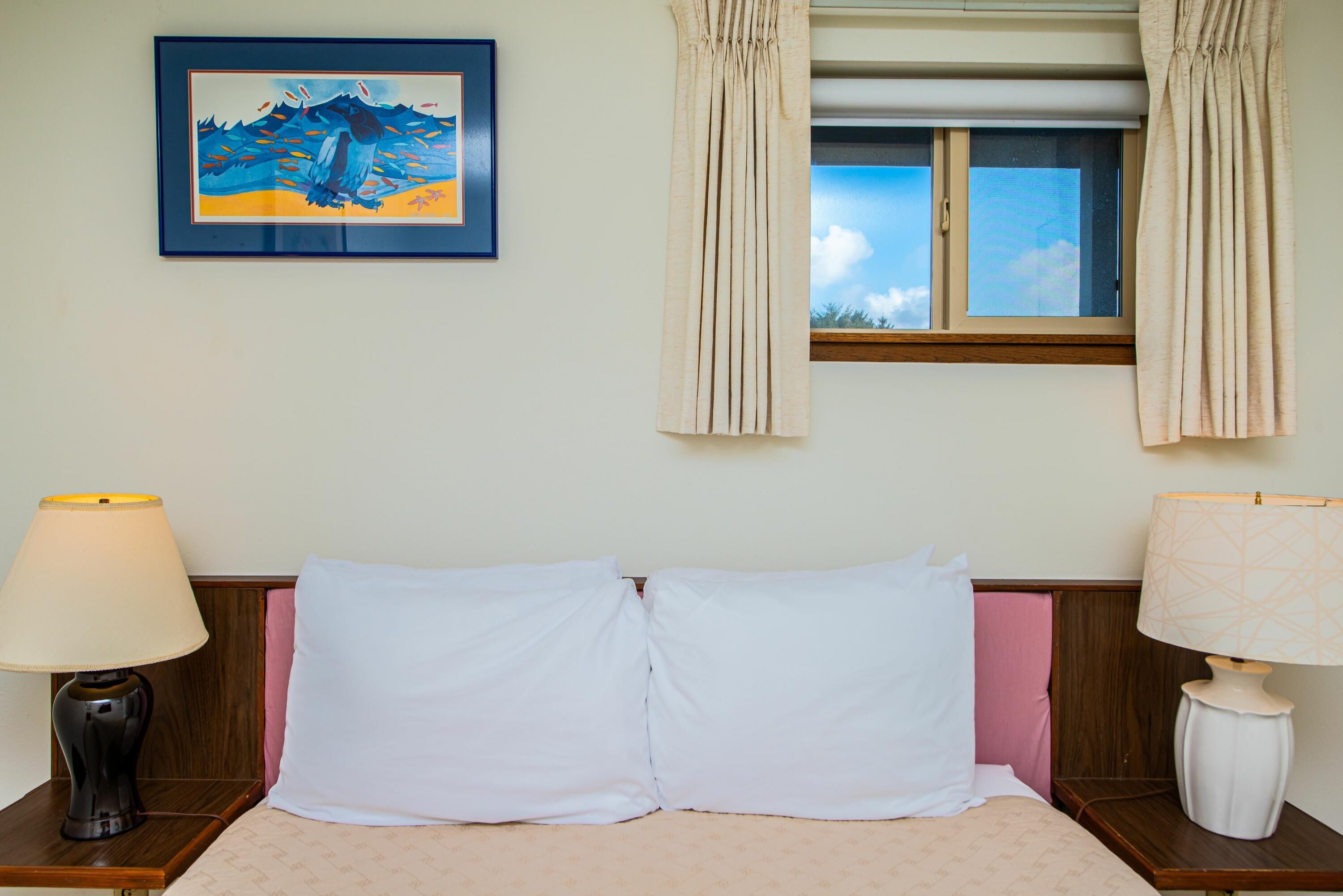 A hotel room with a bed, two bedside lamps, a window with curtains, and a framed painting on the wall.
