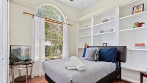 Une chambre à coucher bien rangée, avec un lit, un téléviseur posé sur une table de chevet, une bibliothèque et une fenêtre avec des rideaux.