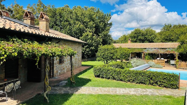 A stone house with a tiled roof, a garden with a swimming pool, and a pergola covered with vines.