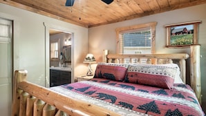 A wooden bed with a patterned bedspread, a nightstand with a lamp, and a window with a view of a house. - Prime Location/Pigeon Forge/3BD/Pet Friendly/Hot Tub/Dollywood/Easy Access (Pigeon Forge)