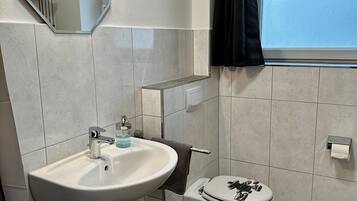 Un baño con lavamanos blanco, espejo, inodoro y ventana con cortina.