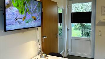Habitació amb un televisor fixat a la paret que mostra ocells, una taula amb un mantell temàtic de cafè, una porta que dona a l'exterior i una finestra amb persianes.