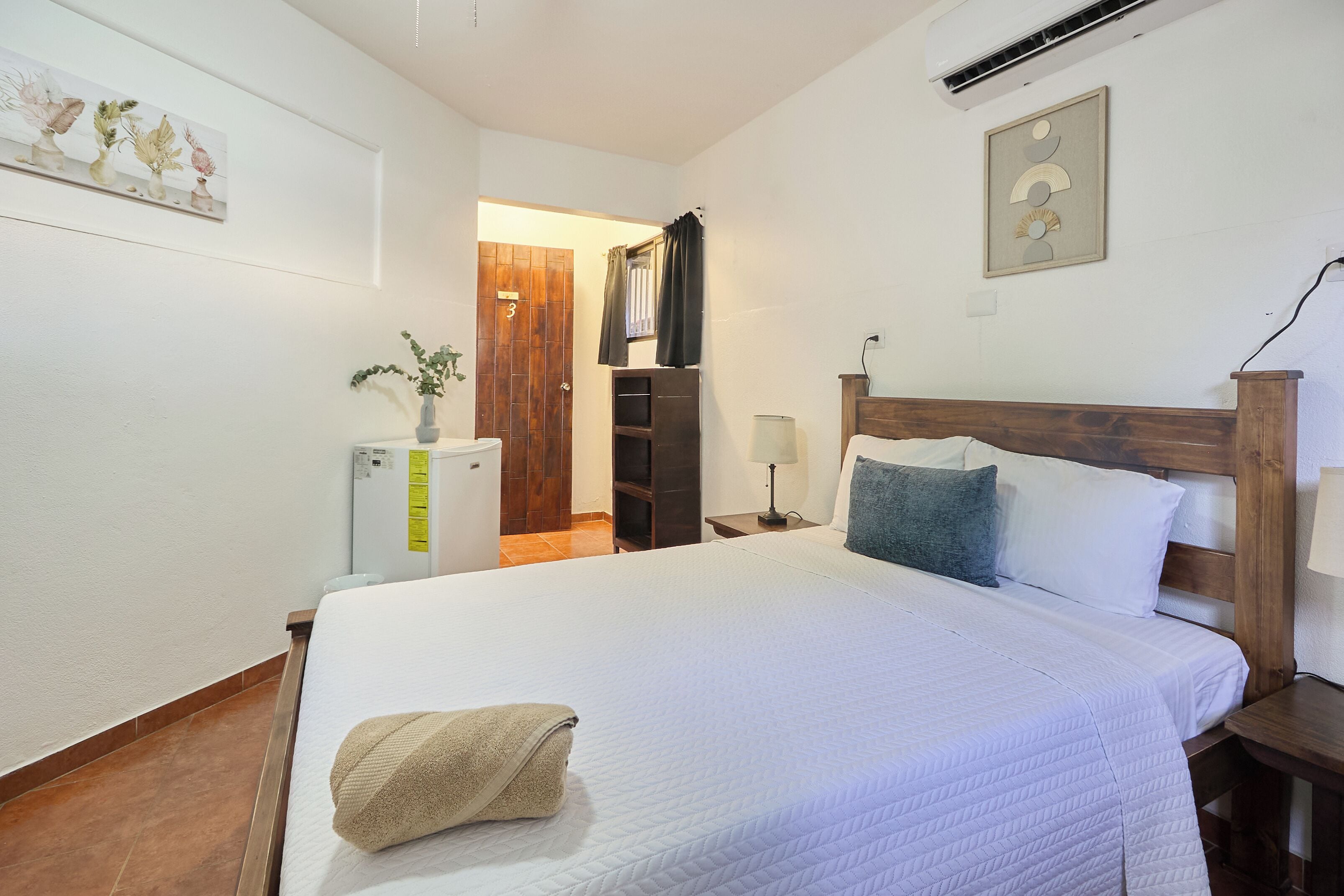 Une chambre d’hôtel avec un grand lit, une tête de lit en bois, une table de chevet, une lampe, une petite table, une plante et une porte donnant sur une autre pièce.