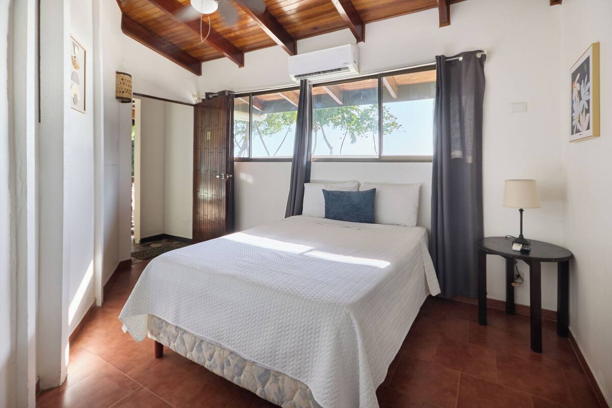 A bedroom with a large bed, a bedside table with a lamp, a window with curtains, and a door leading to another room.