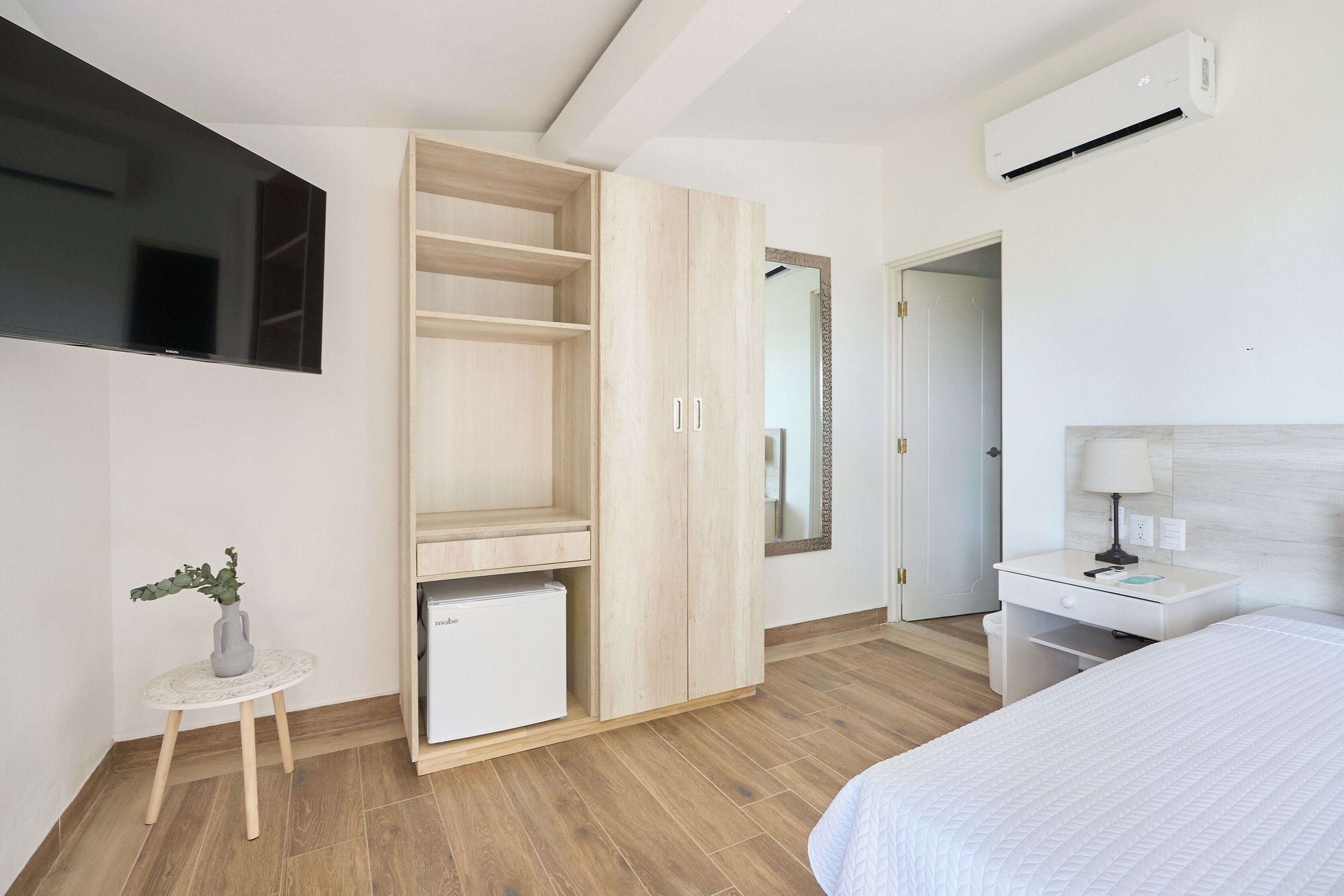 Ein modernes Schlafzimmer mit Bett, Kleiderschrank, kleinem Tisch mit Pflanze, Schreibtisch mit Lampe und an der Wand montiertem Fernseher.