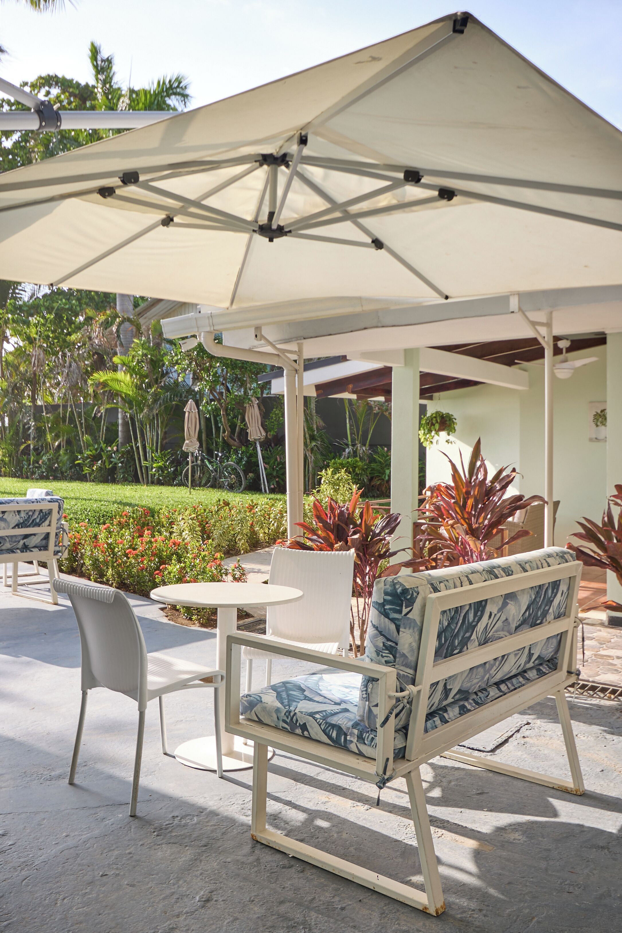 An outdoor seating area with a table and chairs under a large umbrella, surrounded by lush greenery and potted plants.