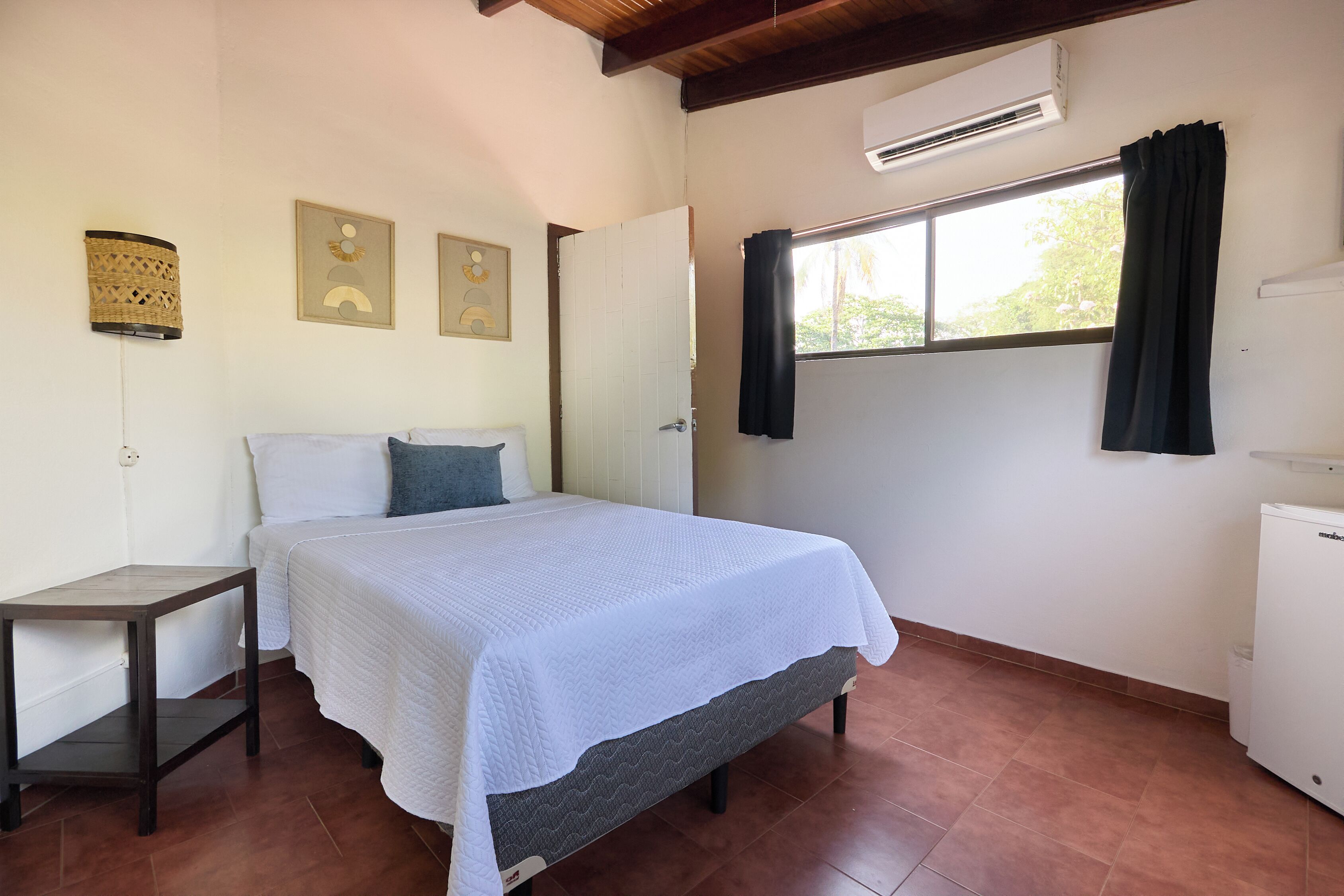 A bedroom with a bed, a nightstand, a window with curtains, and a wall-mounted air conditioner.