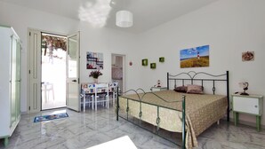 Un dormitorio con cama, mesita de noche y escritorio con sillas. Se ve una puerta que da a otra habitación y una ventana con vistas al exterior.