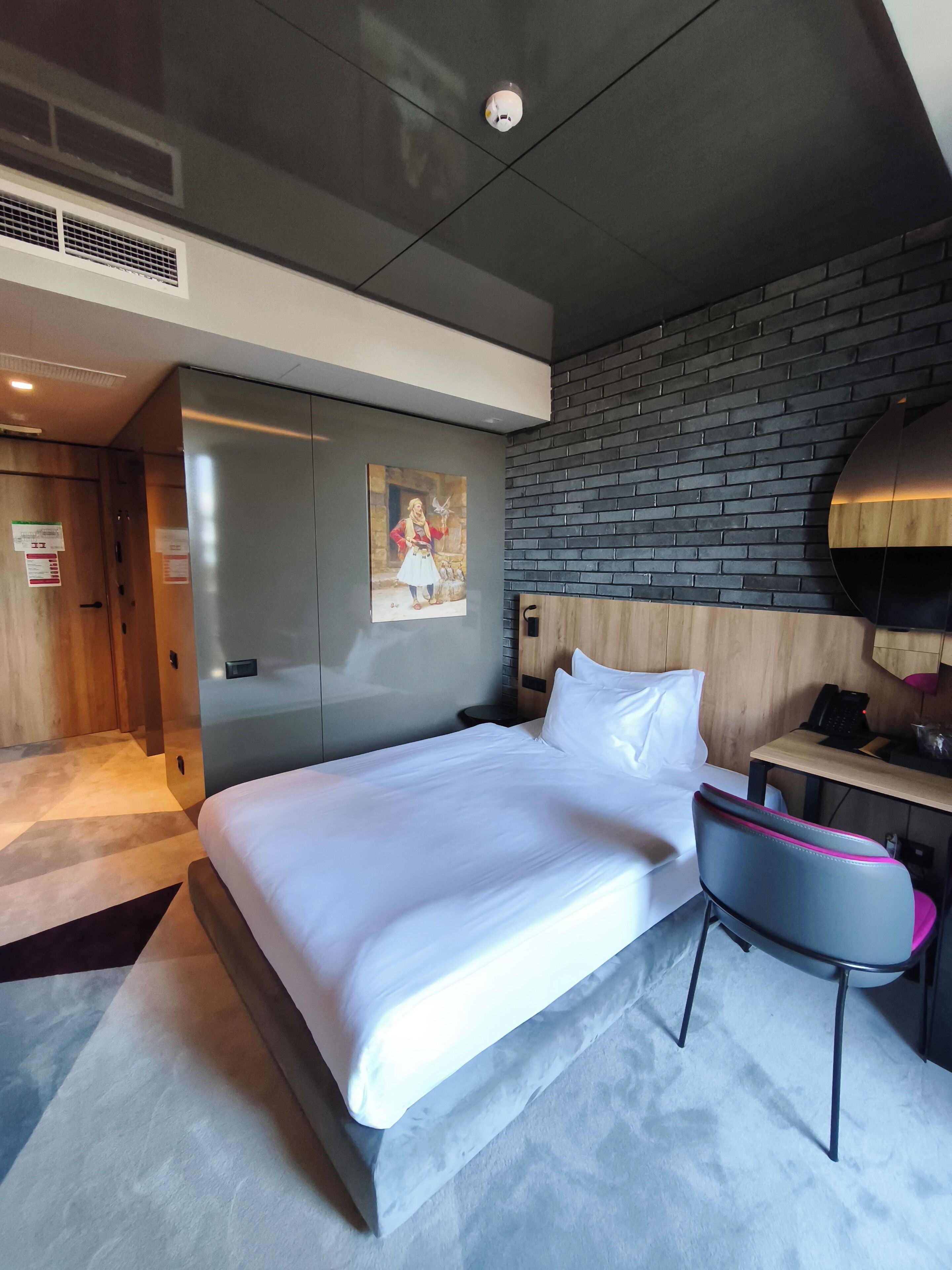 Una habitación de hotel moderna con una cama grande, un cabecero de madera, un escritorio con una silla y una lámpara de pared.