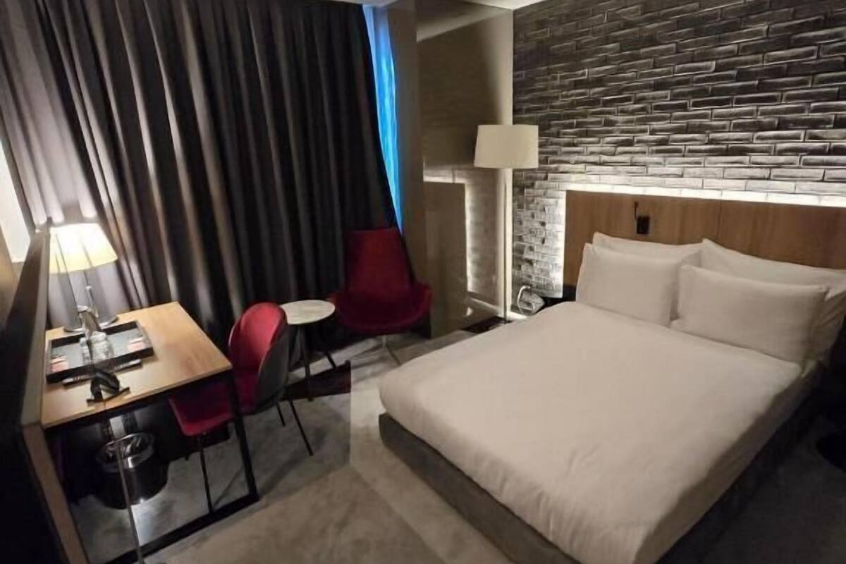 Quarto de hotel moderno com uma cama grande, uma escrivaninha com cadeira, uma mesinha, um abajur e uma parede com padrão de tijolos.