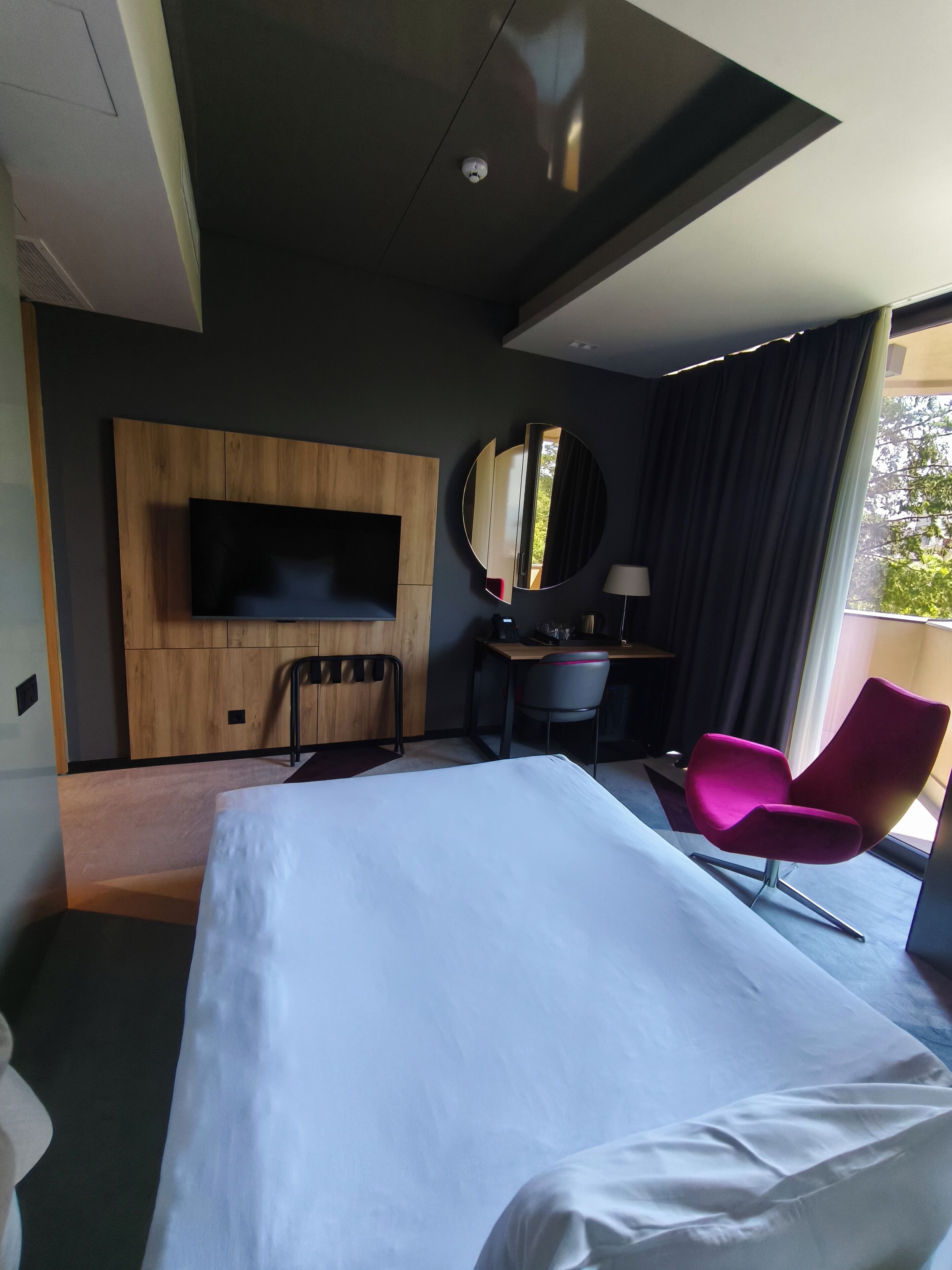 Habitación de hotel moderna con una cama grande, un escritorio, una silla, un televisor montado en la pared y una ventana con cortinas.