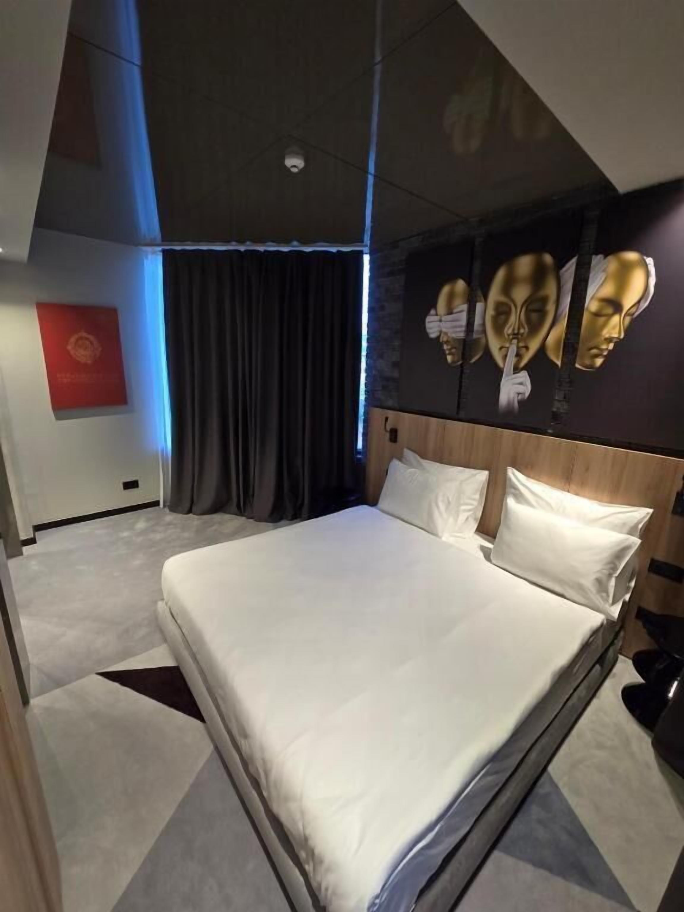 Una habitación de hotel moderna con una cama grande, cortinas oscuras, una cámara de seguridad montada en el techo y una pared con tres máscaras doradas.