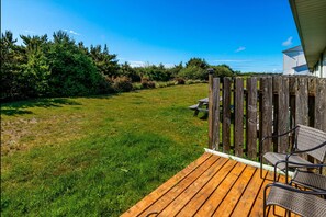 Een houten terras met rieten stoelen en een picknicktafel, met uitzicht op een gazon met bomen en een gebouw op de achtergrond.