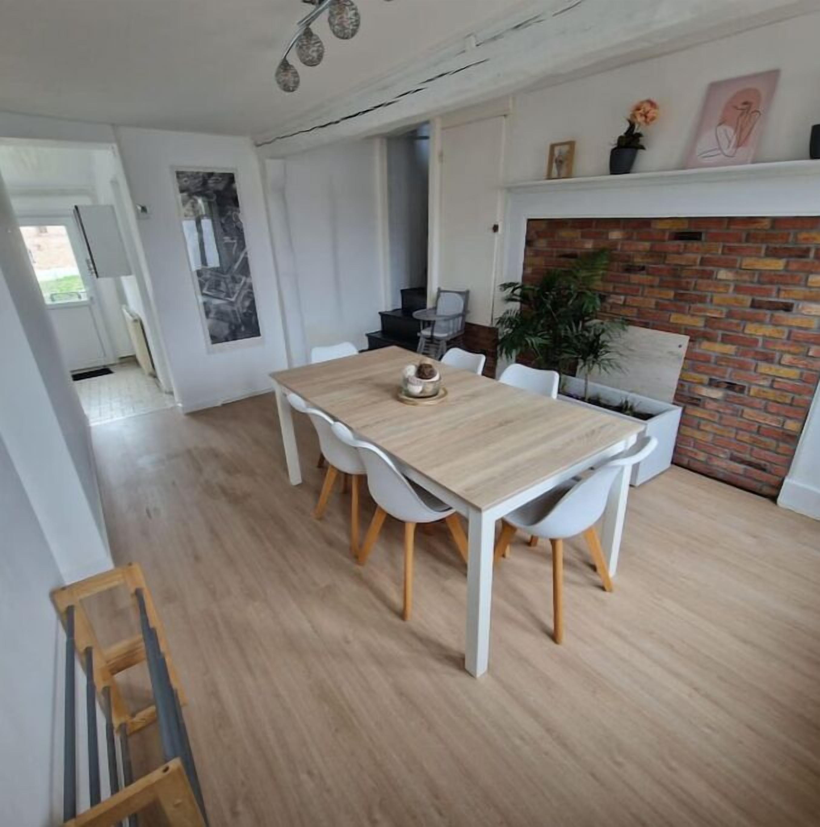 Un comedor con una mesa de madera y sillas blancas, una chimenea de ladrillo y vistas a la cocina.