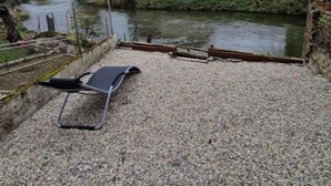 Un patio de grava con una silla metálica, un río y un edificio de fachada blanca y tejado gris.