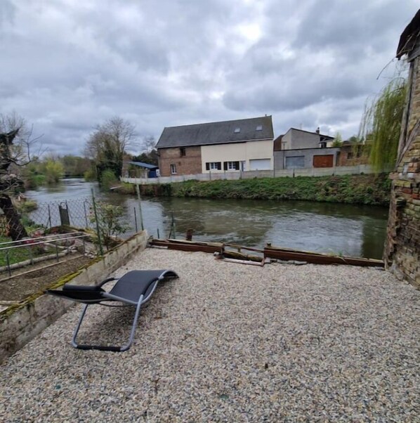 Eine Schotterterrasse mit einem Metallstuhl, einem Fluss und einem Gebäude mit weißer Fassade und grauem Dach.