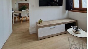 Una sala de estar moderna con un televisor de pantalla plana montado en la pared, un banco de madera con cajones, una mesita con una cesta, y un comedor al fondo.