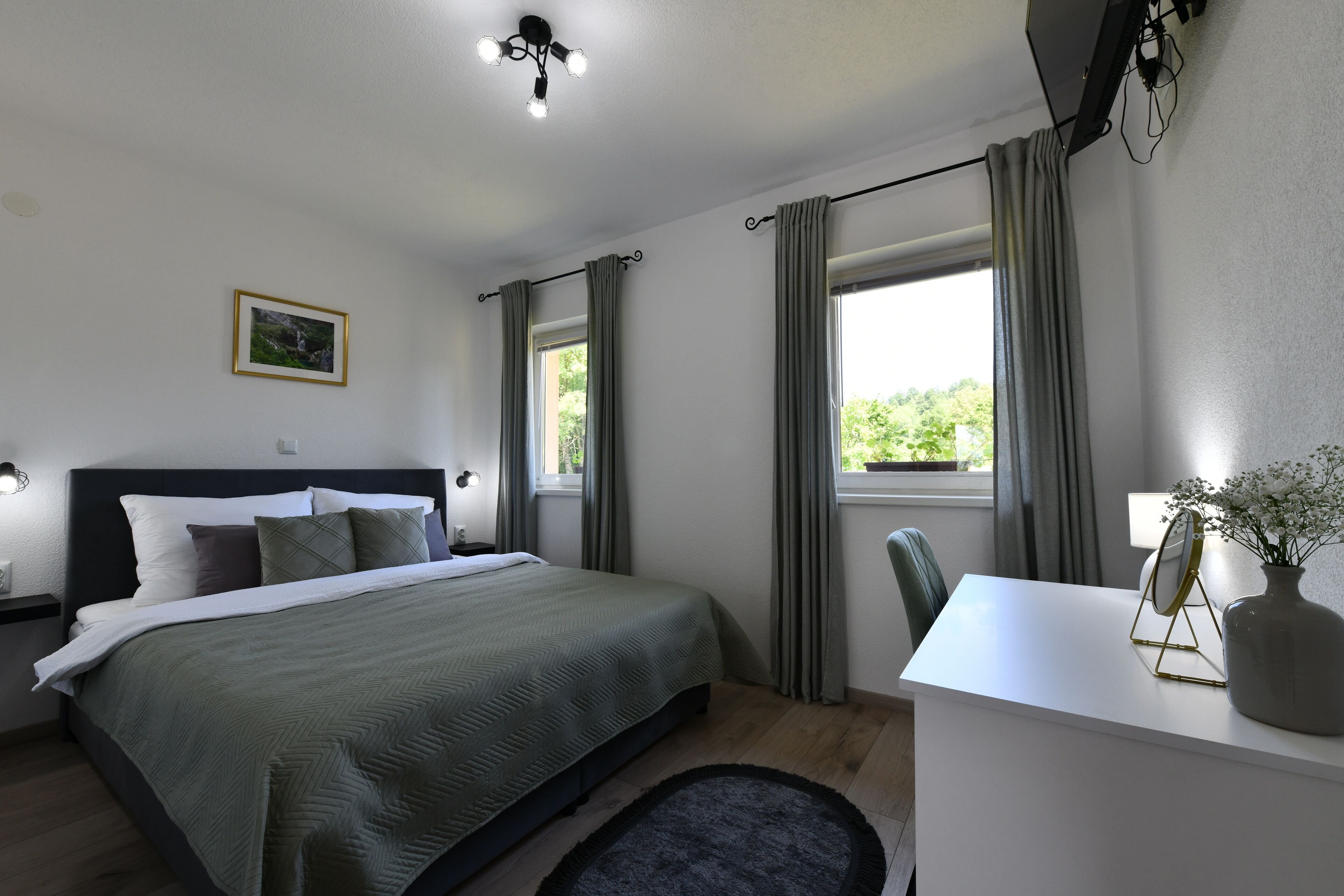 A bedroom with a bed, bedside table, lamp, curtains, and a desk with a vase and mirror.