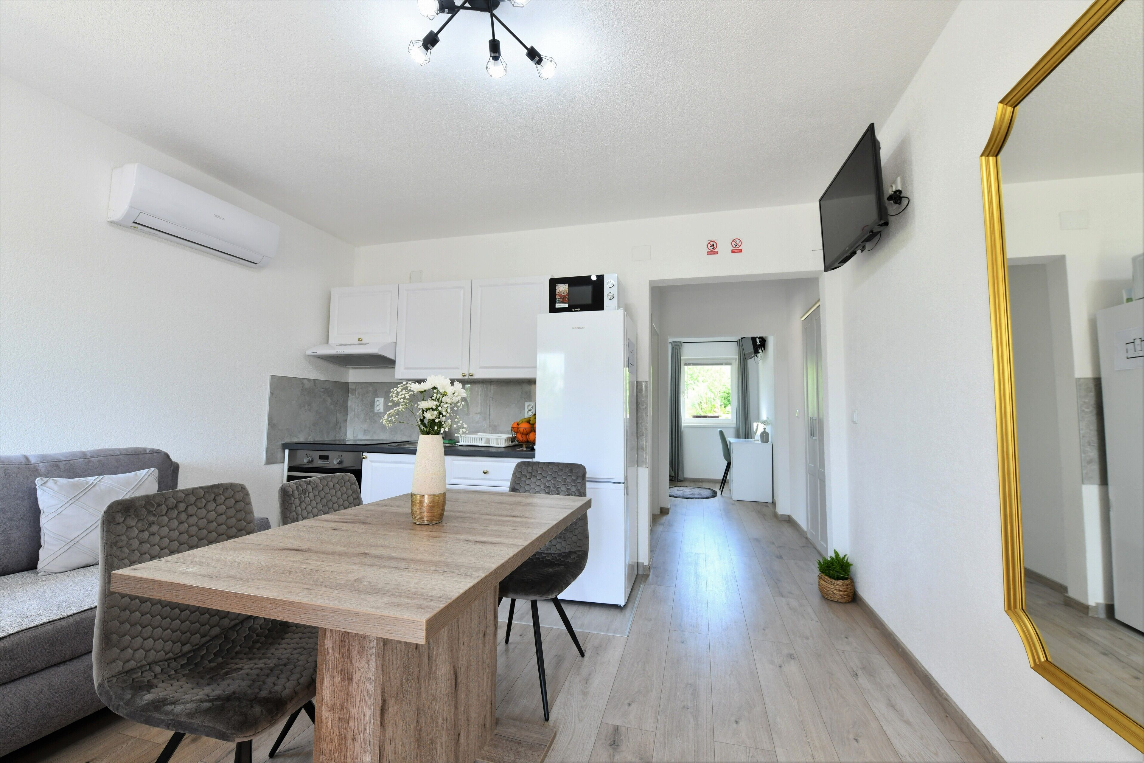 Una cocina moderna con armarios blancos, un microondas y un refrigerador. Hay una mesa de comedor con sillas y un florero con flores. Un espejo grande está en la pared, y hay un equipo de aire acondicionado montado en la pared.