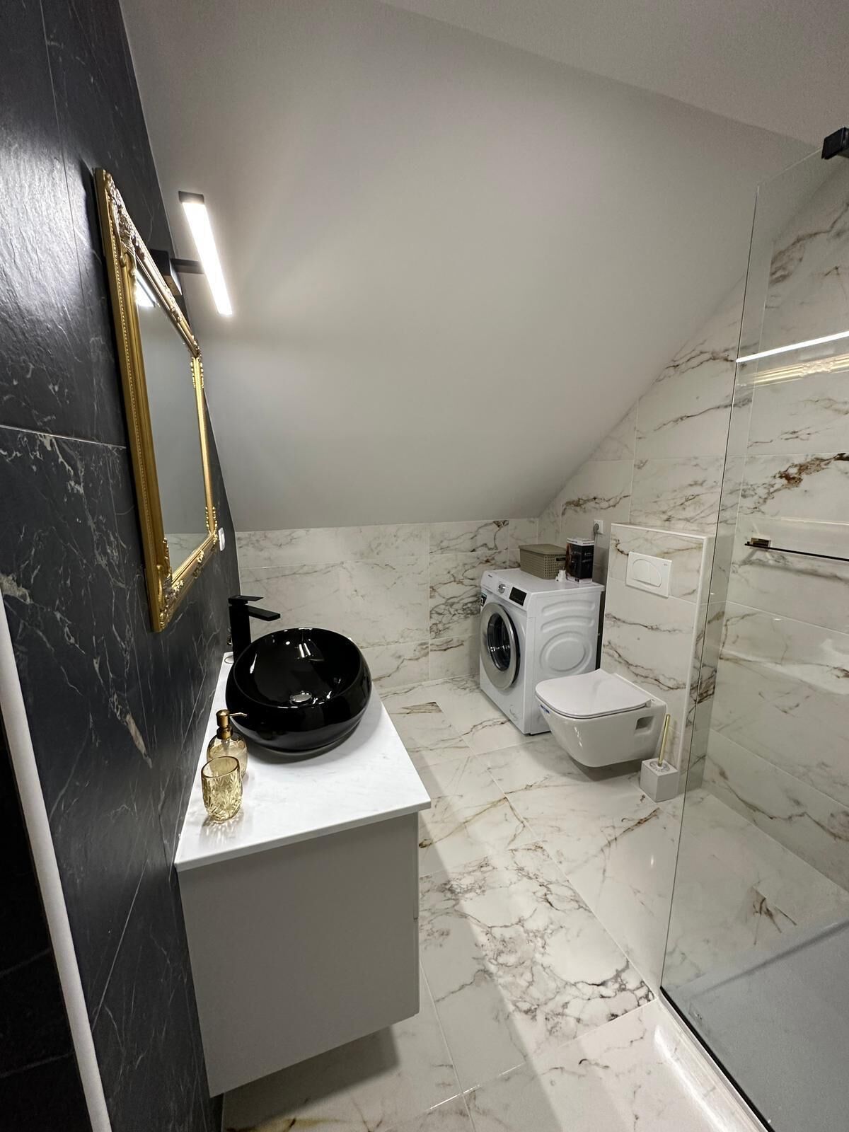 Une salle de bain moderne avec un décor en marbre noir et blanc, un miroir encadré d’or, un lavabo noir et un lave-linge.