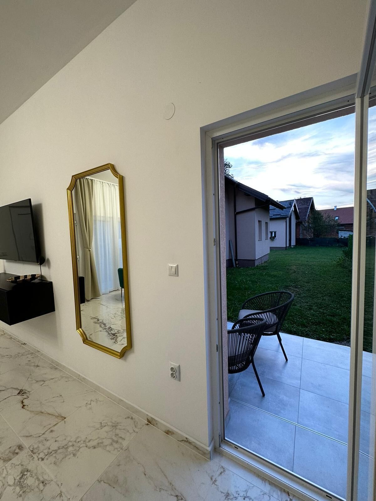 Una sala de estar moderna con un espejo grande, un televisor de pantalla plana y un gran ventanal corredizo que da a un patio con una silla y una mesa pequeña.