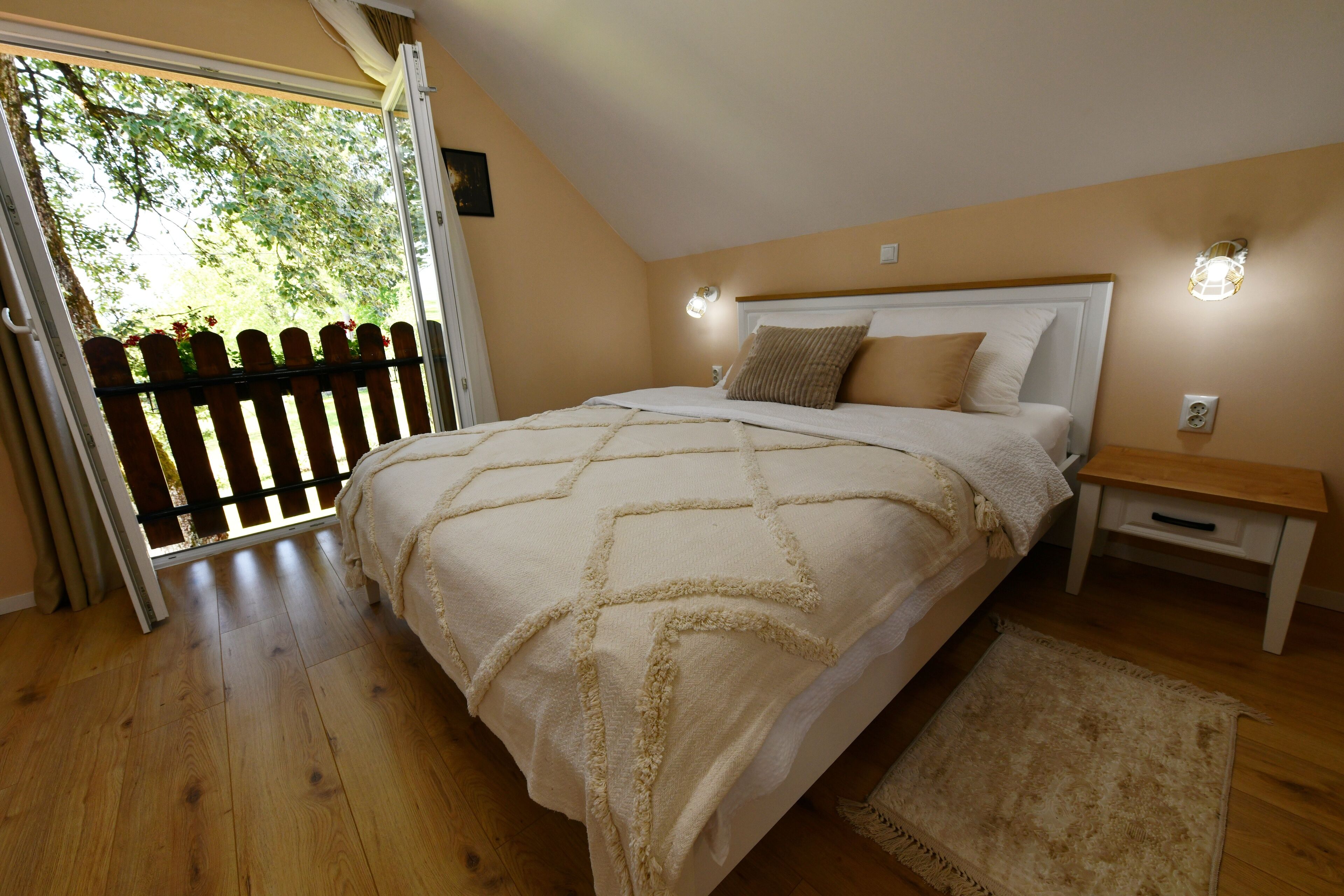 Une chambre avec un plancher en bois, un lit recouvert d’une couette beige, une table de chevet et une vue sur un espace extérieur, visible à travers une porte vitrée, cet espace étant délimité par une clôture en bois.