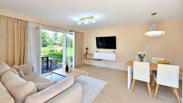 A modern living room with a sofa, a dining table with chairs, a TV mounted on the wall, and a sliding glass door leading to a garden.