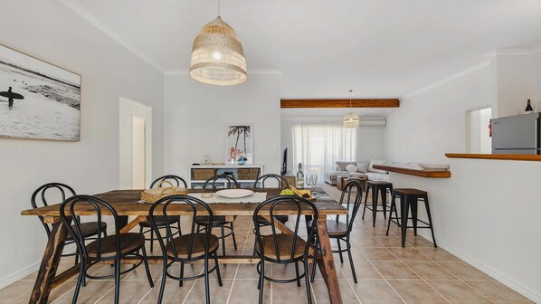 A spacious dining area with a wooden table and chairs, a hanging light fixture, and a kitchen area with a refrigerator and a shelf. - Light & Bright Pet Friendly Beach Side Haven (Dawesville)