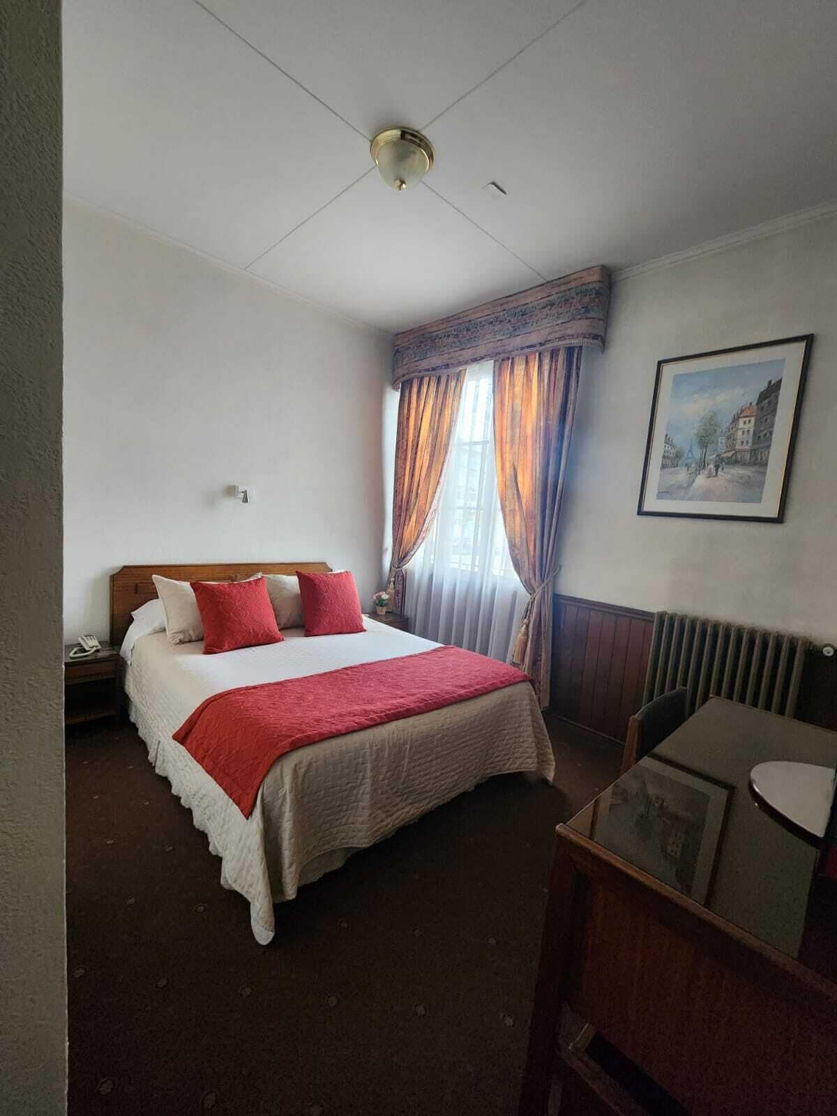 Una habitación de hotel con cama, escritorio, silla, radiador y un cuadro en la pared.