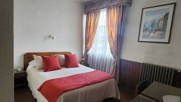Una habitación de hotel con cama, escritorio, silla, radiador y un cuadro en la pared.