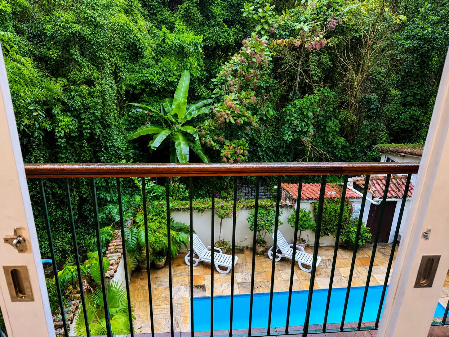 Piscina cercada por vegetação exuberante, um terraço com azulejos e cadeiras de descanso brancas.