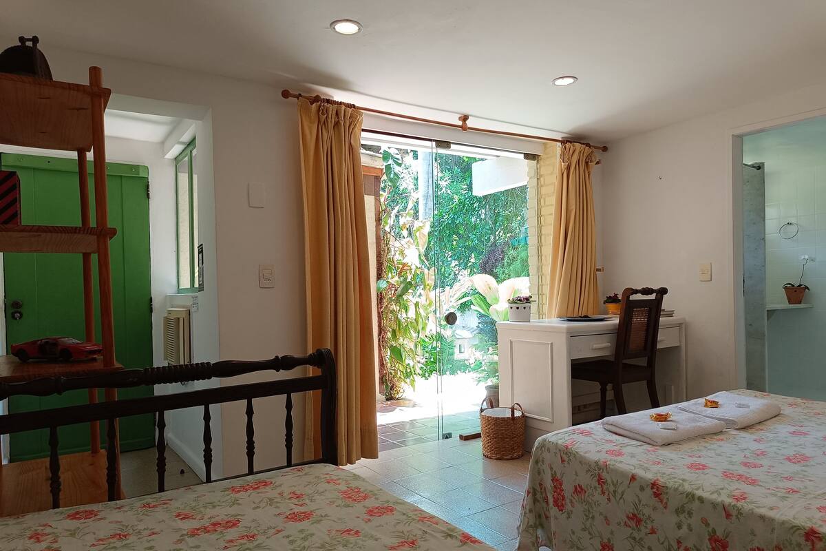 A room with two beds, a desk, a chair, a window with curtains, and a view of greenery outside.